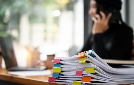 Pile of papers and in the background someone on the phone looking at a laptop