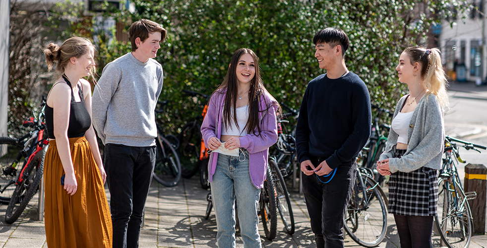 Group of five students stood outside in a huddle smiling and talking to one another