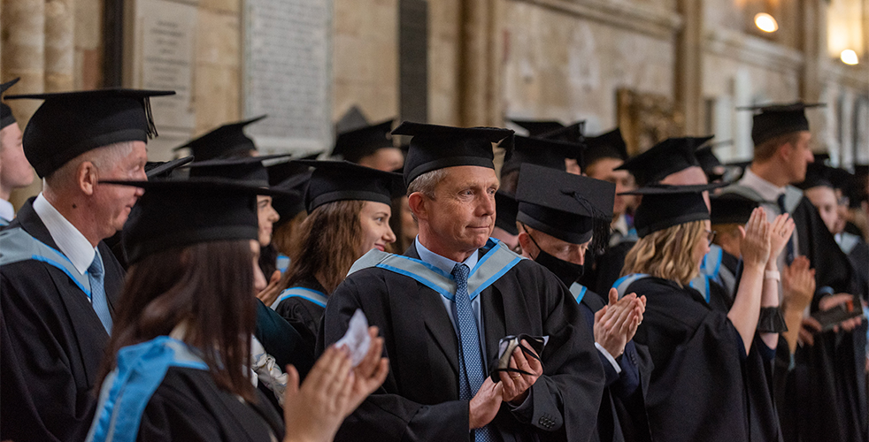 Adult learners in cap and gown clapping at college graduation ceremony in Exeter Cathedral
