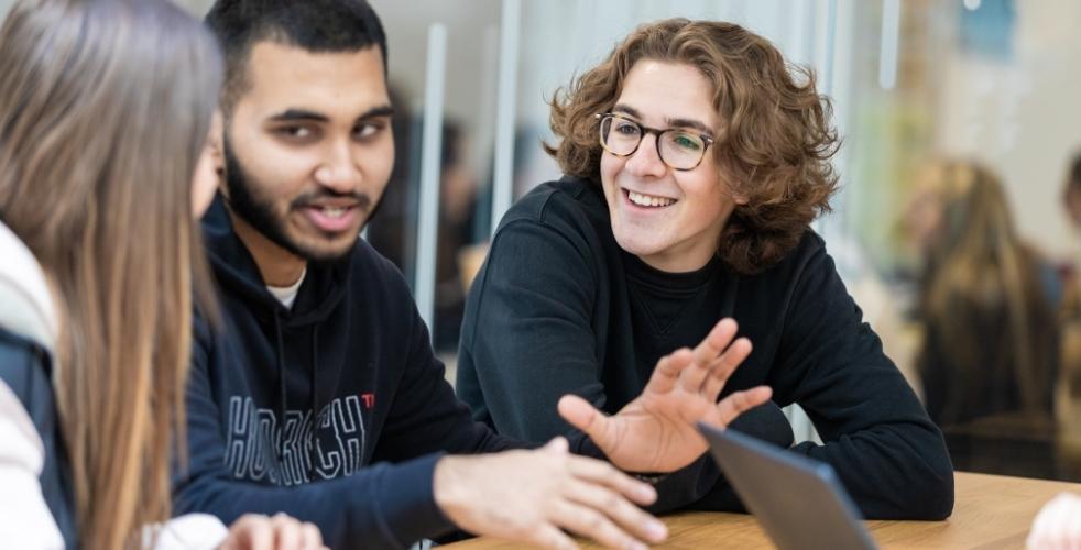 Three students sat at a table talking with one leaning forward