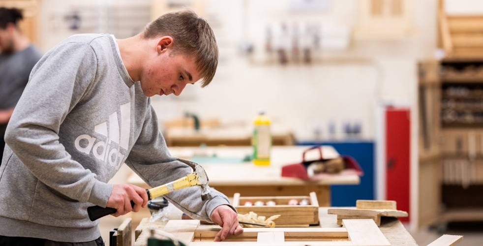 Male student holding a hammer over the top of a nail in a grey adidas jumper