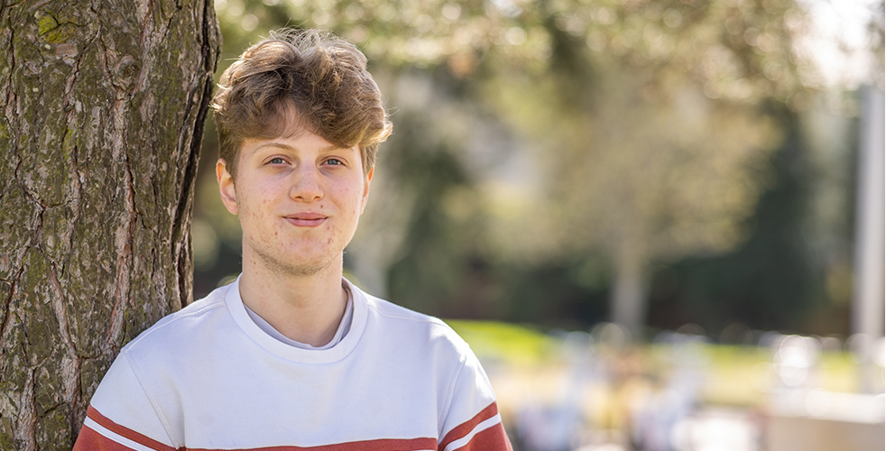 Male Student portrait smiling at the camera leaning on a tree wearing a white and red stripped sweatshirt