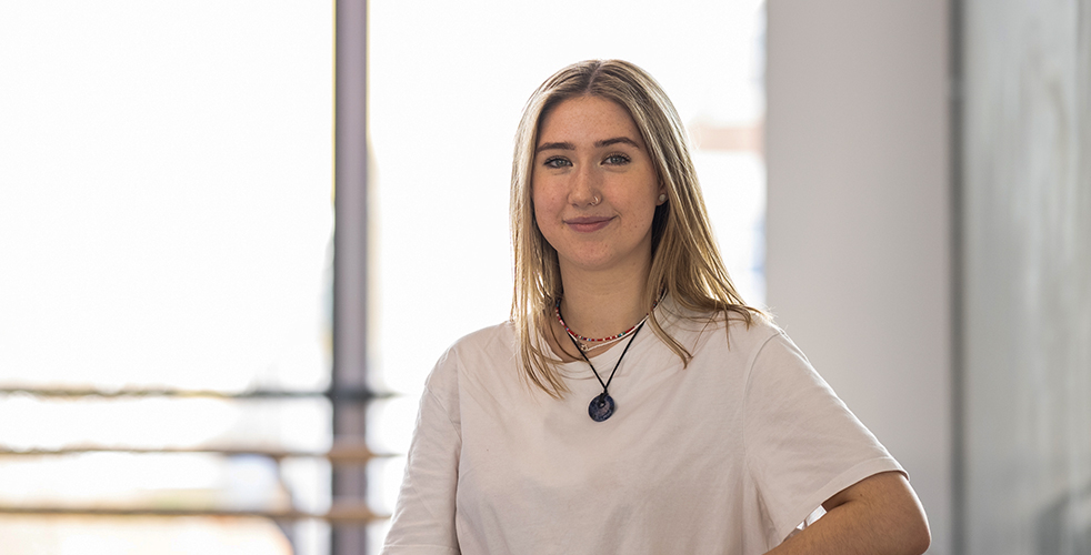 Female Student portrait smiling at the camera wearing a white top leaning on a wooden bar