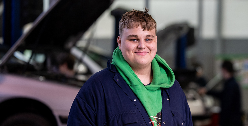 Male portrait of a student smiling at the camera in a green hoodie and blue overalls with cars in the background