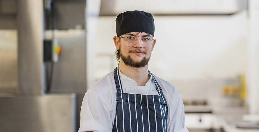 Male Student portrait smiling at the camera stood in a kitchen in his chef whites and a black and white stripped apron