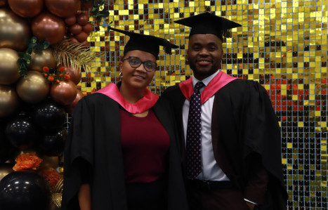 Two students smile at the camera in their graduation robes.