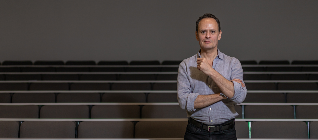 A male stands in a lecture theatre in chinos and a shirt.