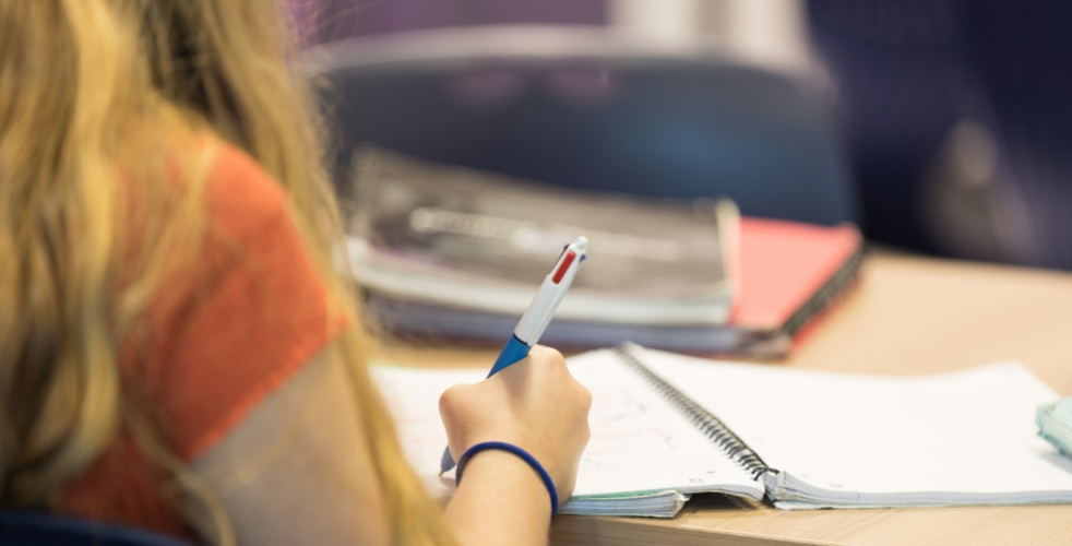 Image of a student's shoulder with focus on the pen they are holding, writing on some paper in a notebook on a desk