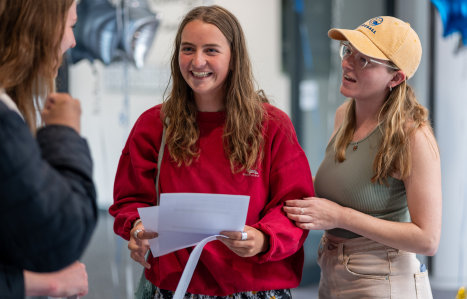 A student wearing red holds her results and smiles at the camera.