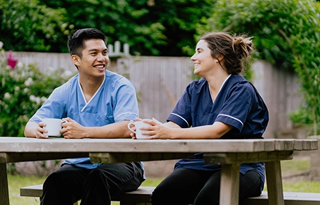 Two colleagues in scrubs sat on a bench smiling and talking