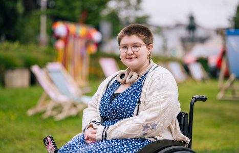 A young women sits in a wheelchair and smiles at the camera. She wears a blue dress.