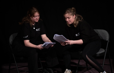 Two students sat on chairs on a dark stage holding scripts interacting with each other