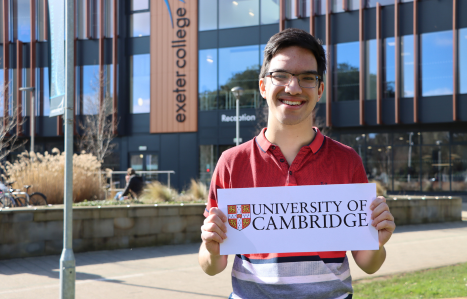 A young man smiles holding a University of Cambridge sign