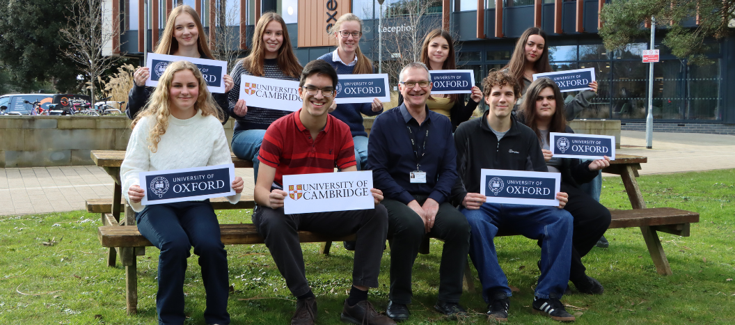 Male and female students sit on a bench holding an Oxford or Cambridge sign