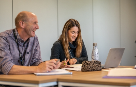 two students smiling at a desk with a laptop on it in class