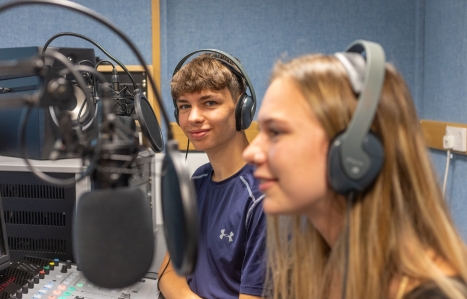 Two students talking into mics at a radio station with one smiling at the camera