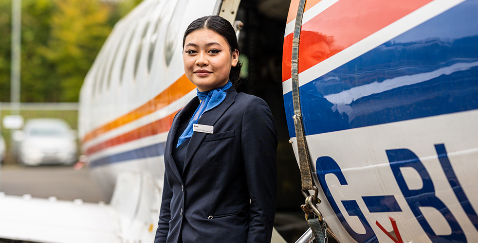 Female Student portrait smiling at the camera wearing a blazer with a blue neck tie around her neck stood in front of a plane