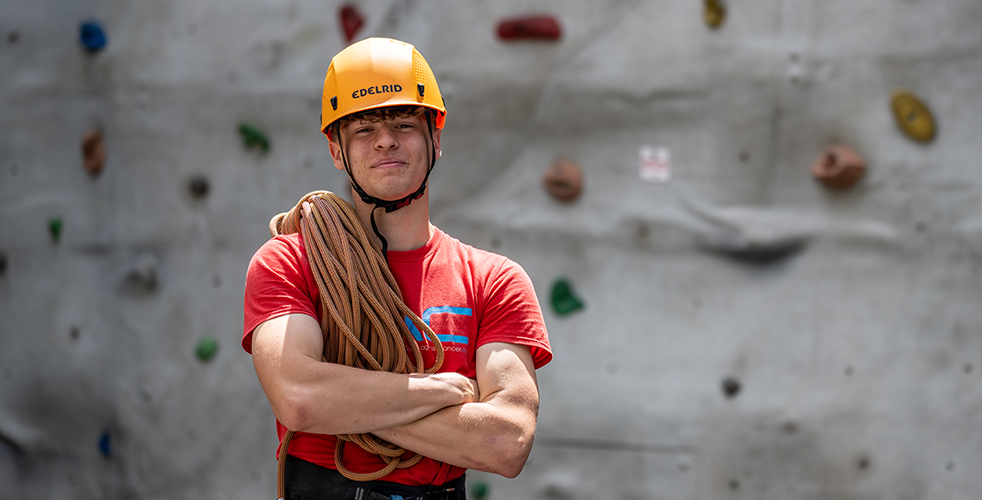 Male Student portrait smiling at the camera wearing a red top with a hard hat and climbing rope over his shoulder