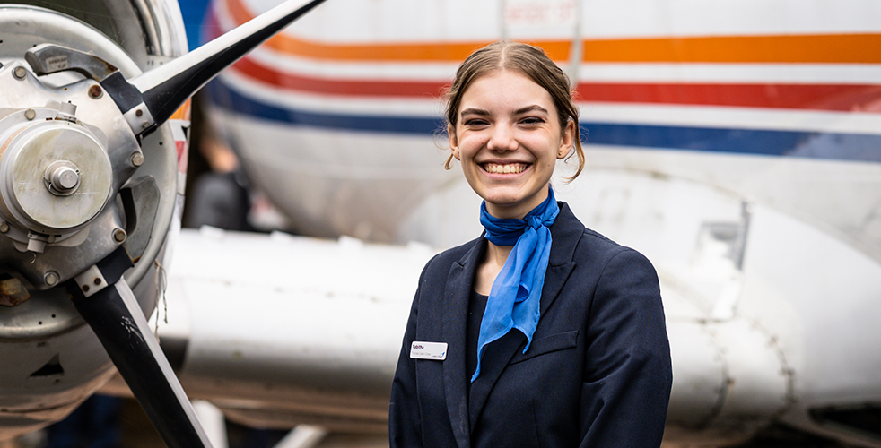 Female Student portrait smiling at the camera wearing a blazer with a blue neck tie around her neck stood in front of a plane
