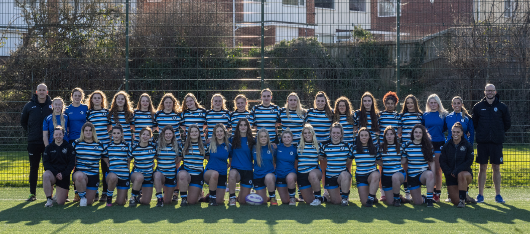 Exeter College's Women's Rugby team in their kits knelling on the side of the pitch for a group/team photo