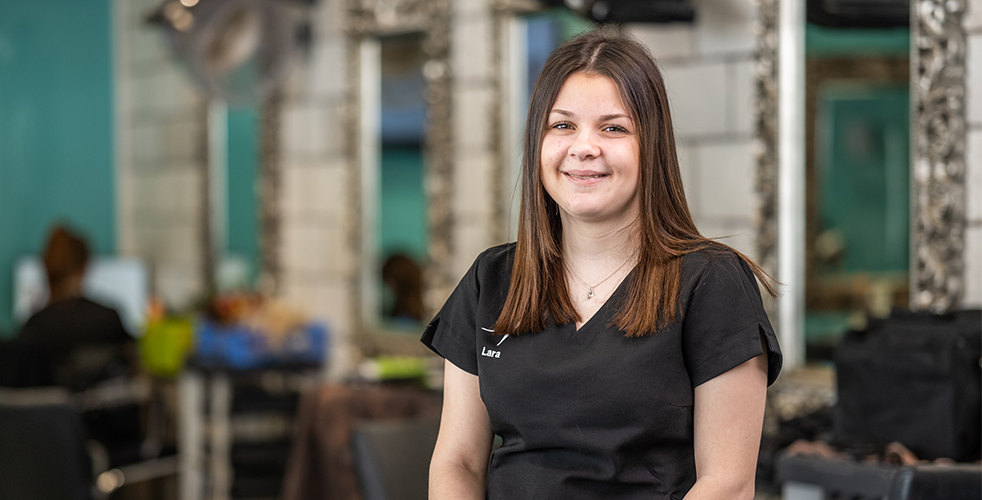Female Student portrait smiling at the camera wearing a black work top with their name on standing in the hair and beauty salon