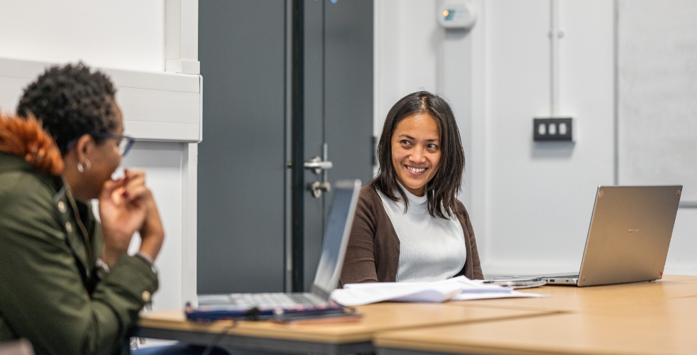 Two students sat at a desk on their laptops smiling at each other