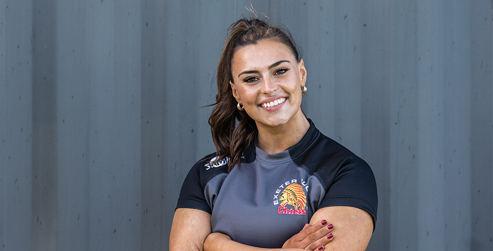 female Exeter Chiefs rugby student smiling at the camera in front of a grey wall