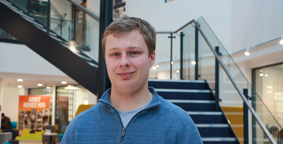 Male student smiling at the camera with short blonde/brunette hair in a blue quarter zip jumper