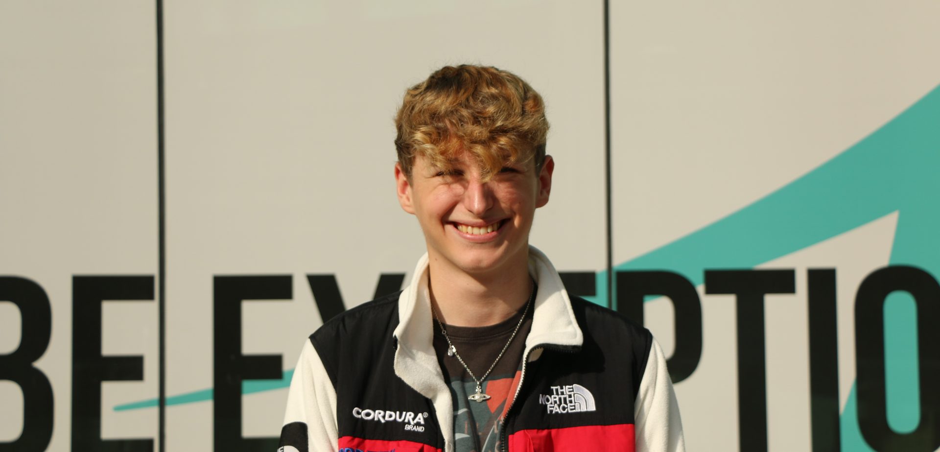 Student smiling at the camera wearing a white black and red north face jacket in front of the be exceptional