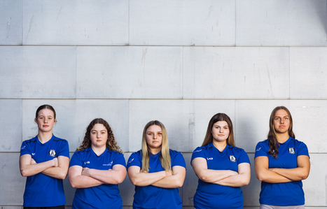Group of five girls stood in front of a grey wall with their arms crossed staring at the camera
