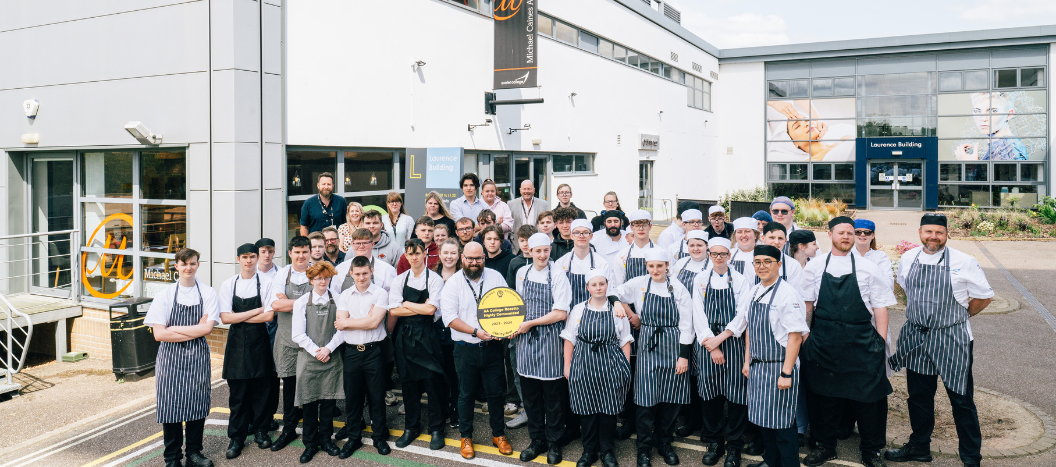 A group of catering students standing in chef whites and black and white aprons in front of the Michael Caines @34 building.