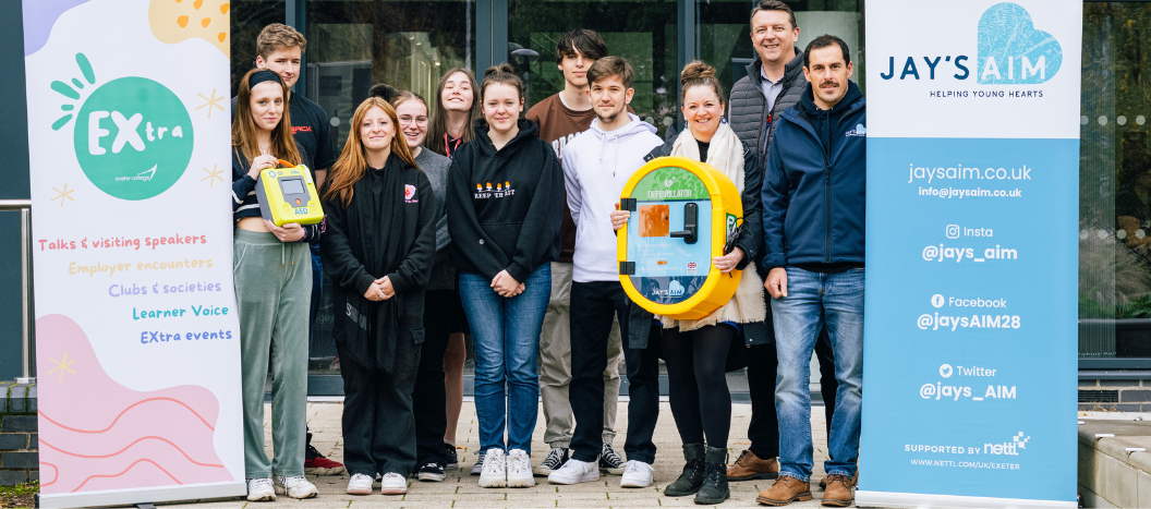 Groups of students stood between two Banners (Extra and Jay's Aim) holding a defibrillator