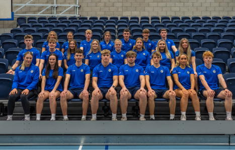 A group of sports students sit on benches in a sports hall.