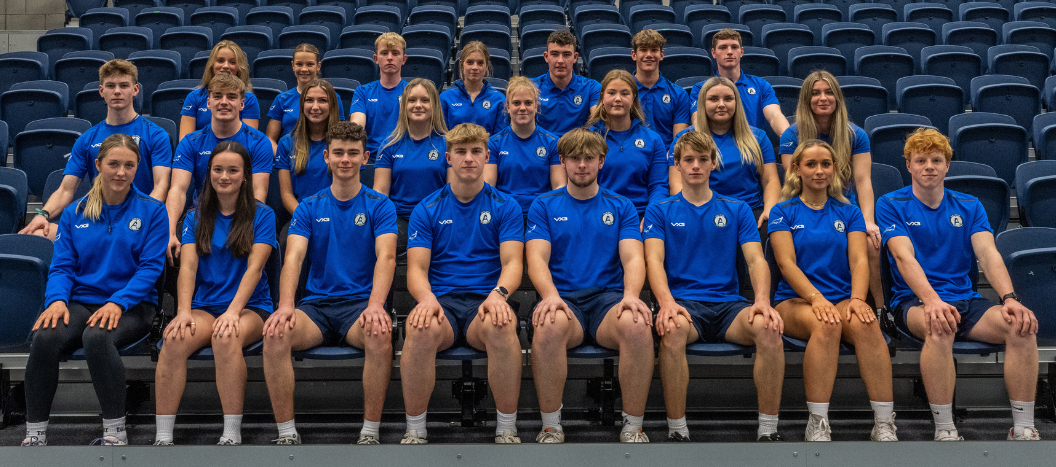A group of sports students sit on benches in a sports hall.