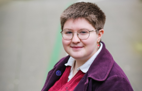 A girl with short hair and circular glasses in a purple jacket smiles at the camera