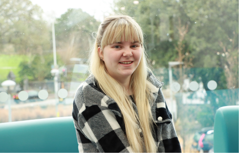 A girl with blonde hair smiles at the camera wearing a black and white chequered shirt.
