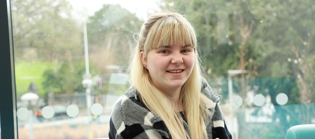A girl with blonde hair smiles at the camera wearing a black and white chequered shirt.