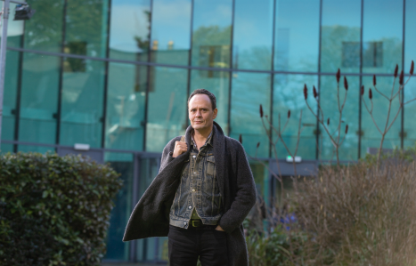 A male stands outside the IoT building in a jacket, shirt and chinos