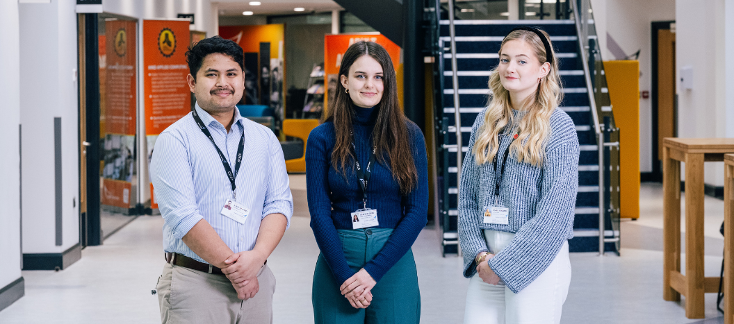 Two female and one male apprentices stand in a line in the foyer of a building.