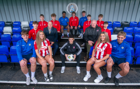 A group of female and male football players sit on some pitch side seating.