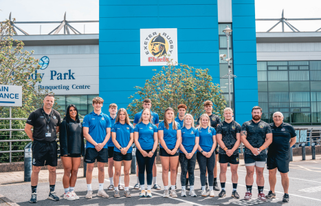 A group of female and male rugby players and coaches stand in front of Sandy Park.