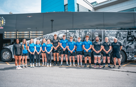 A group of female and male rugby players and coaches stand in front of the Exeter Chiefs coach at Sandy Park.