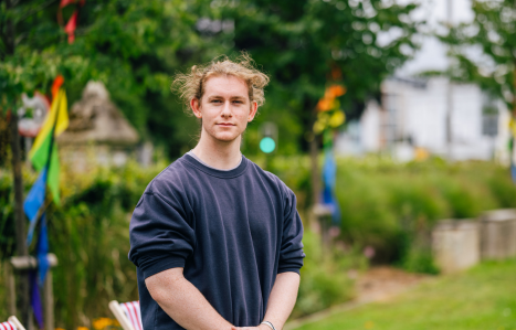 A young male student in a grey polo jumper smiles at the camera.