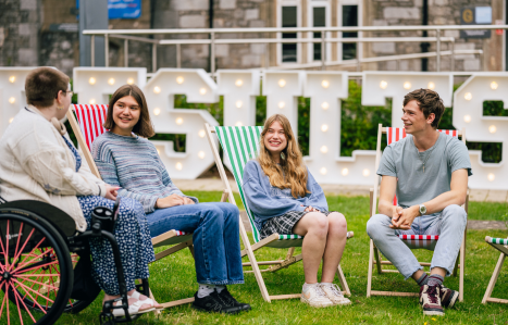 A group of friends sit on deckchairs smiling on campus.