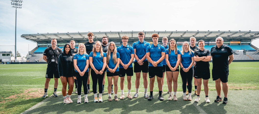 A group of male and female rugby players and coaches stand on the pitch at Sandy Park