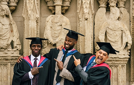 Three students in their graduation gowns stood outside the cathedral smiling with arms around one another