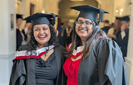 Two adult students in their graduation gowns smiling with arms round each other