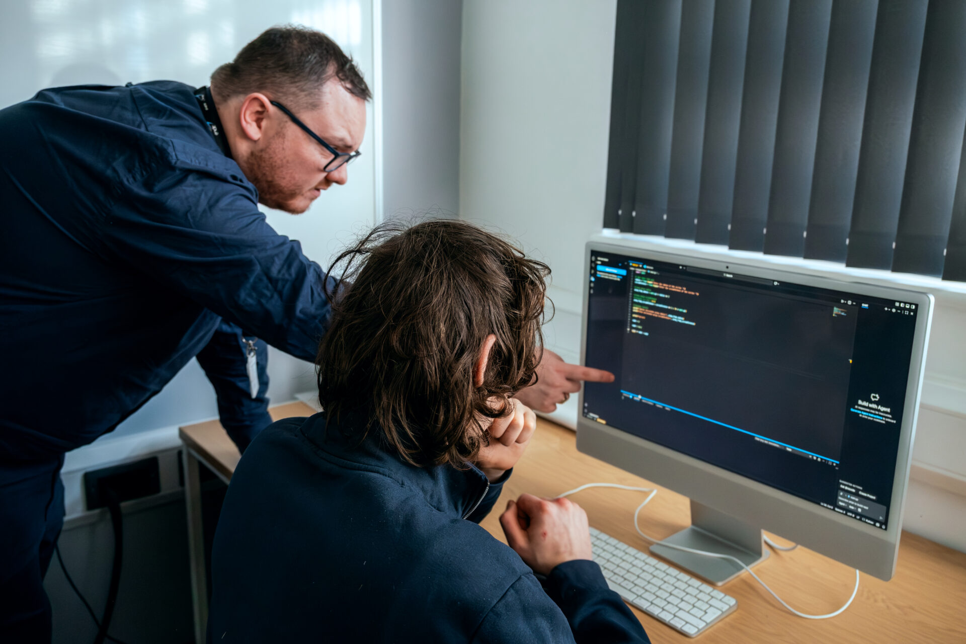 student reviewing code on mac computer with teacher stood beside them pointing at screen