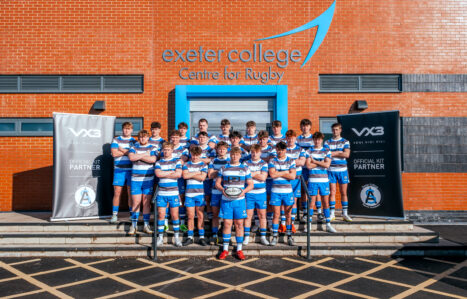 Male Rugby Team in their kits stood on steps out side brick building with the Exeter College Logo on