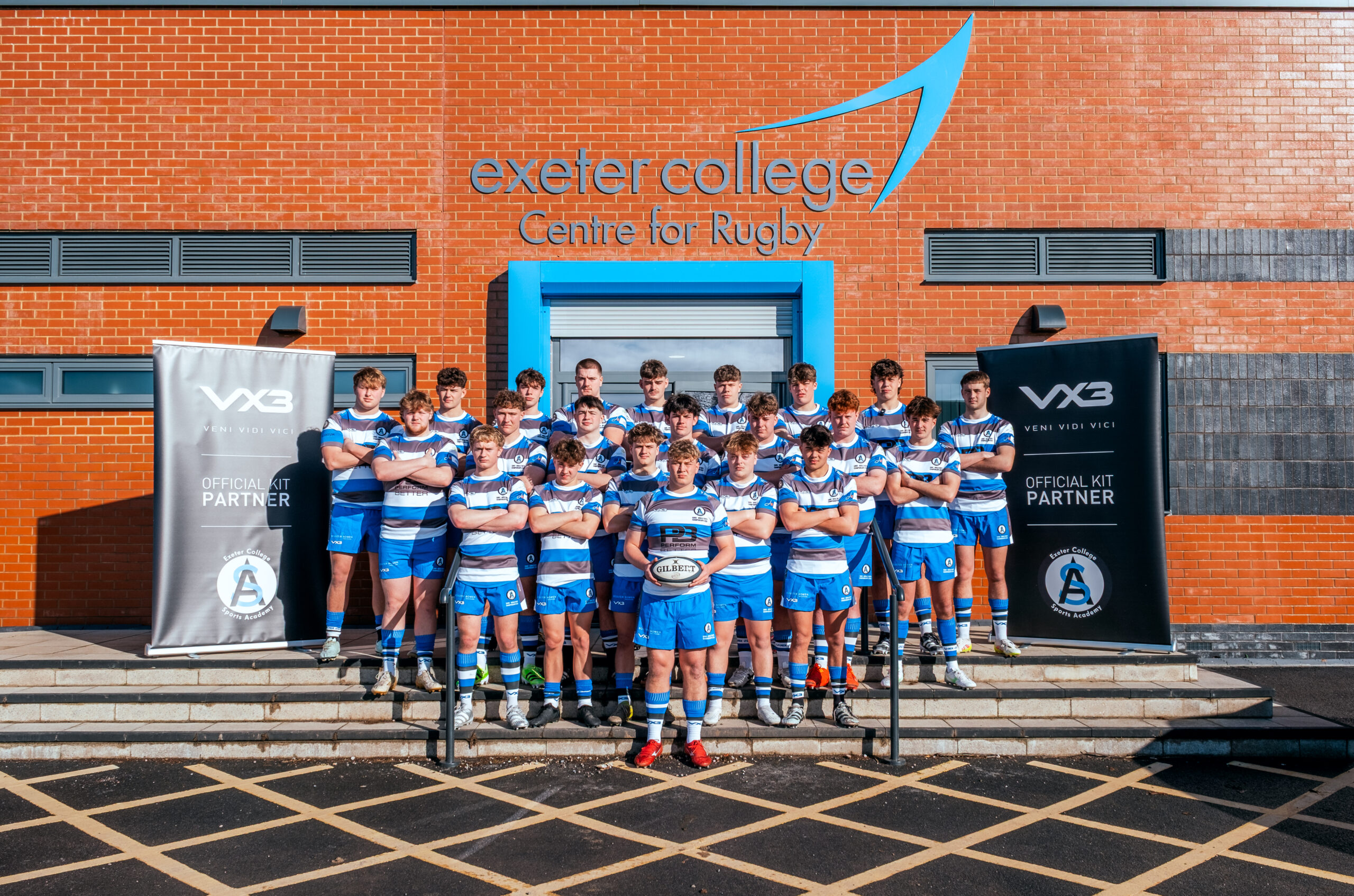 Male Rugby Team in their kits stood on steps out side brick building with the Exeter College Logo on
